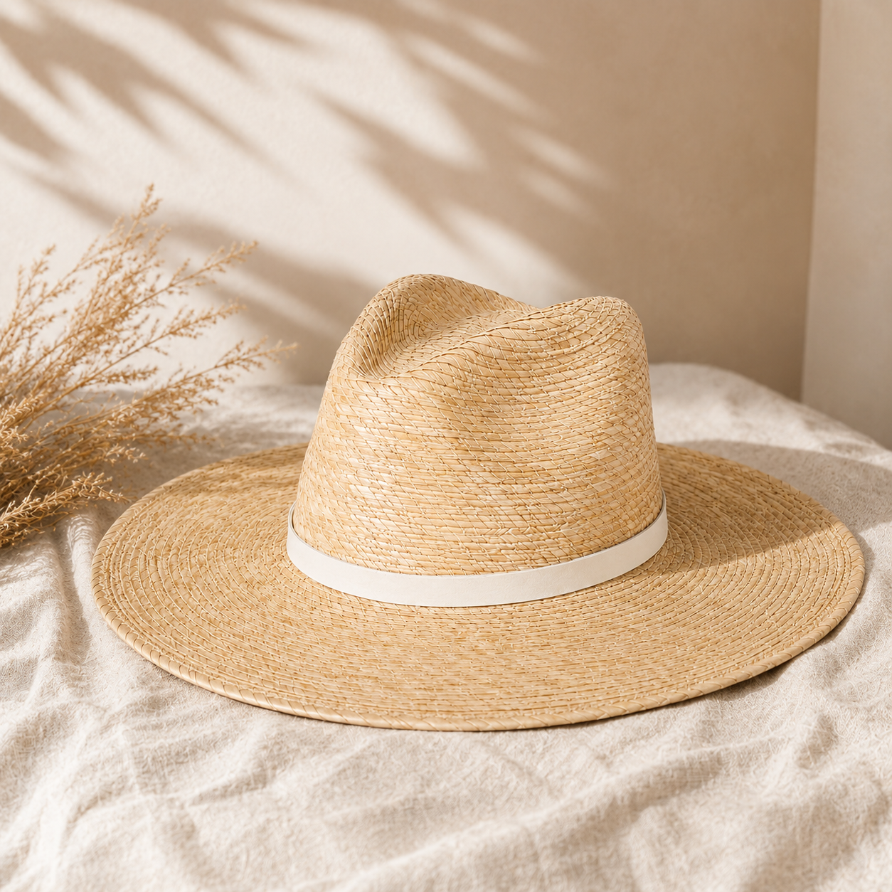 Beige straw hat with a white band on a beige surface with dried plants.
