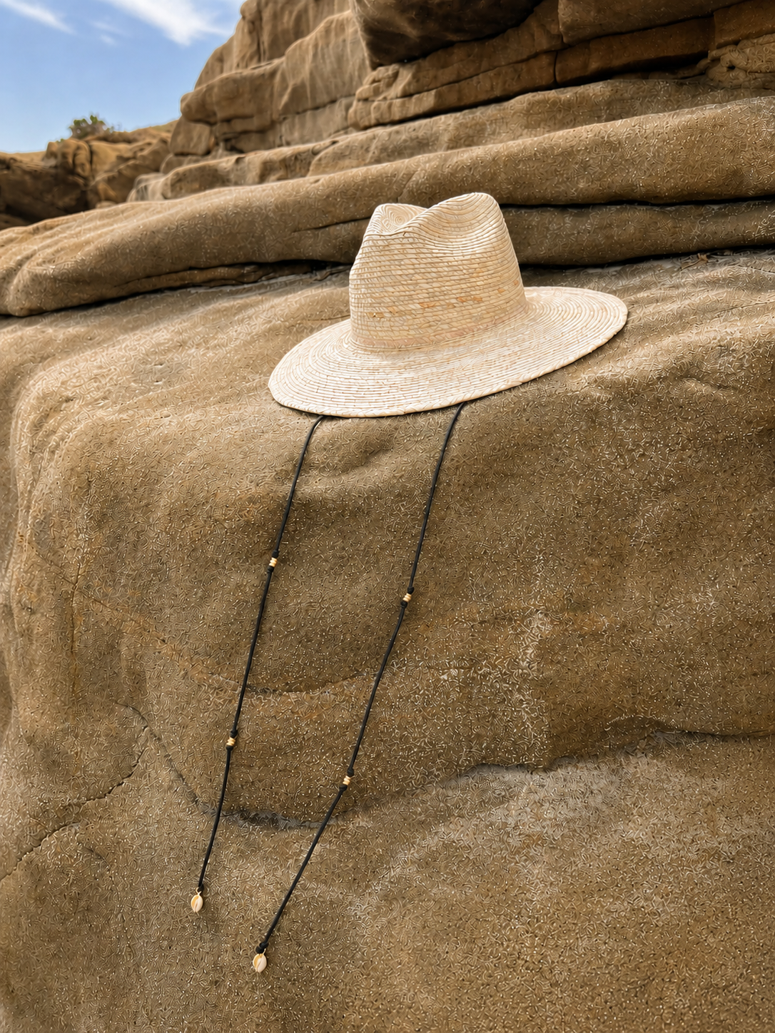 A light beige woven sun hat with a black cord necklace draped over the brim, resting on a large tan rock outdoors.