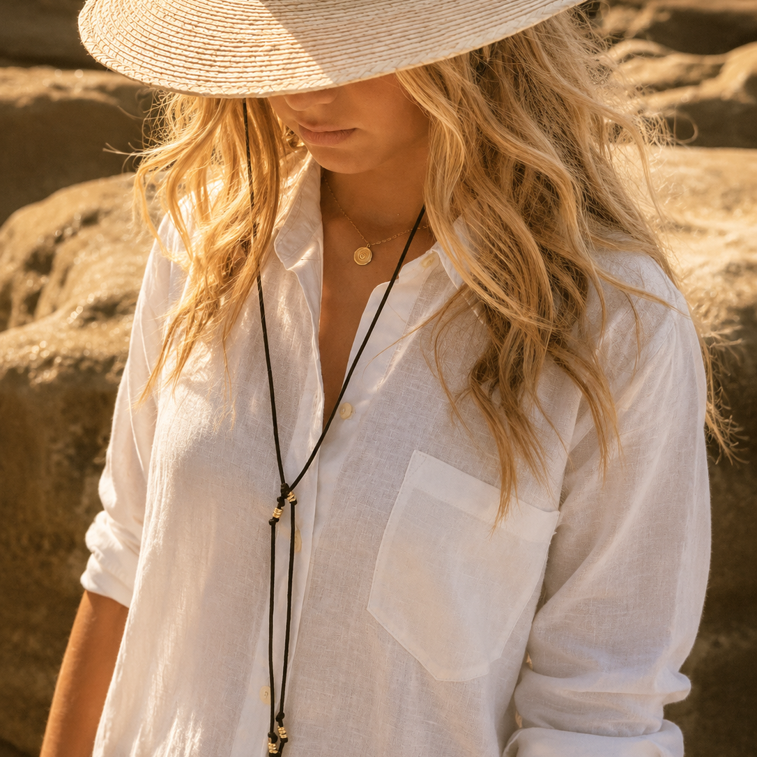 A woman wearing a wide-brimmed straw hat, a white linen button-up shirt, and a long black cord necklace, photographed outdoors against a warm sandy background.