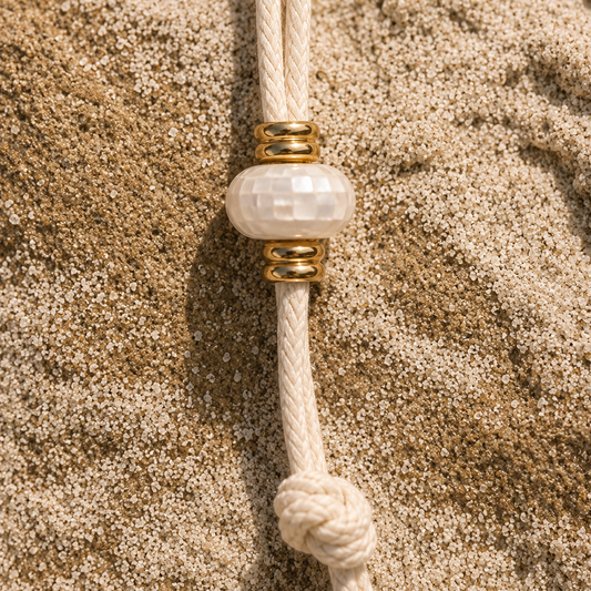 A close-up of a cream-colored braided cord bracelet or necklace with a faceted white bead and gold spacer rings, resting on a textured sandy background.