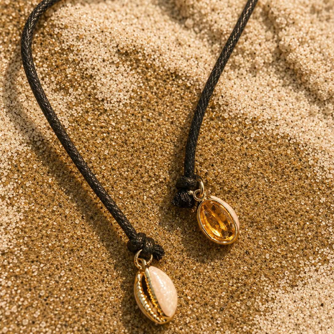 Two cowrie shell necklaces on black cords, one with a gold-toned shell and one with a white shell, resting on a sandy beach background.