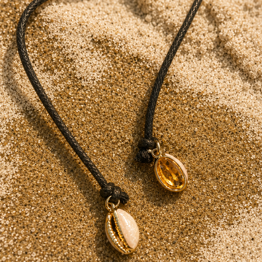 Two cowrie shell necklaces on black cords, one with a gold-toned shell and one with a white shell, resting on a sandy beach background.