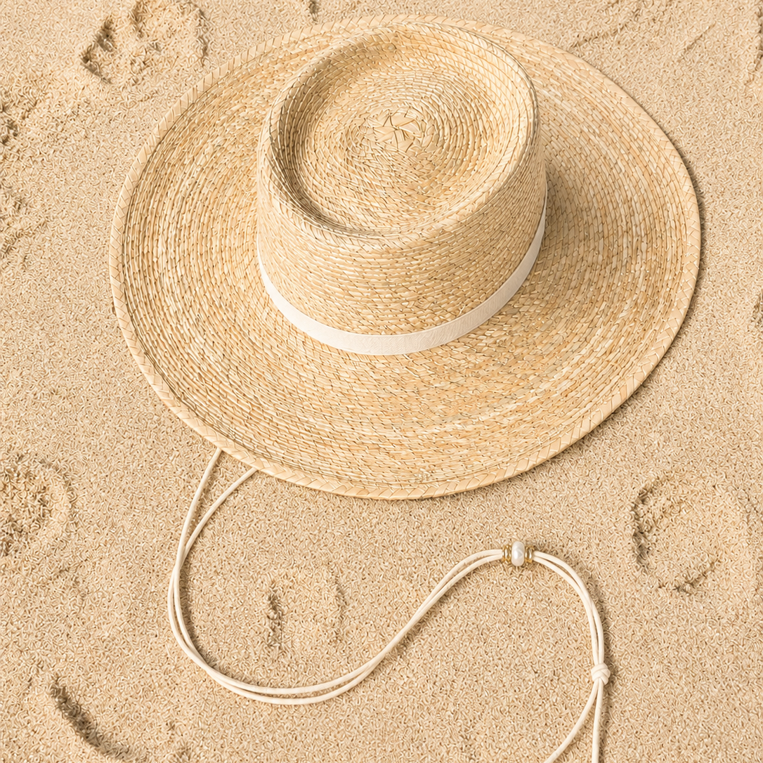 A natural straw sun hat with a white band and matching chin strap, laid flat on sandy ground with footprints visible around it.