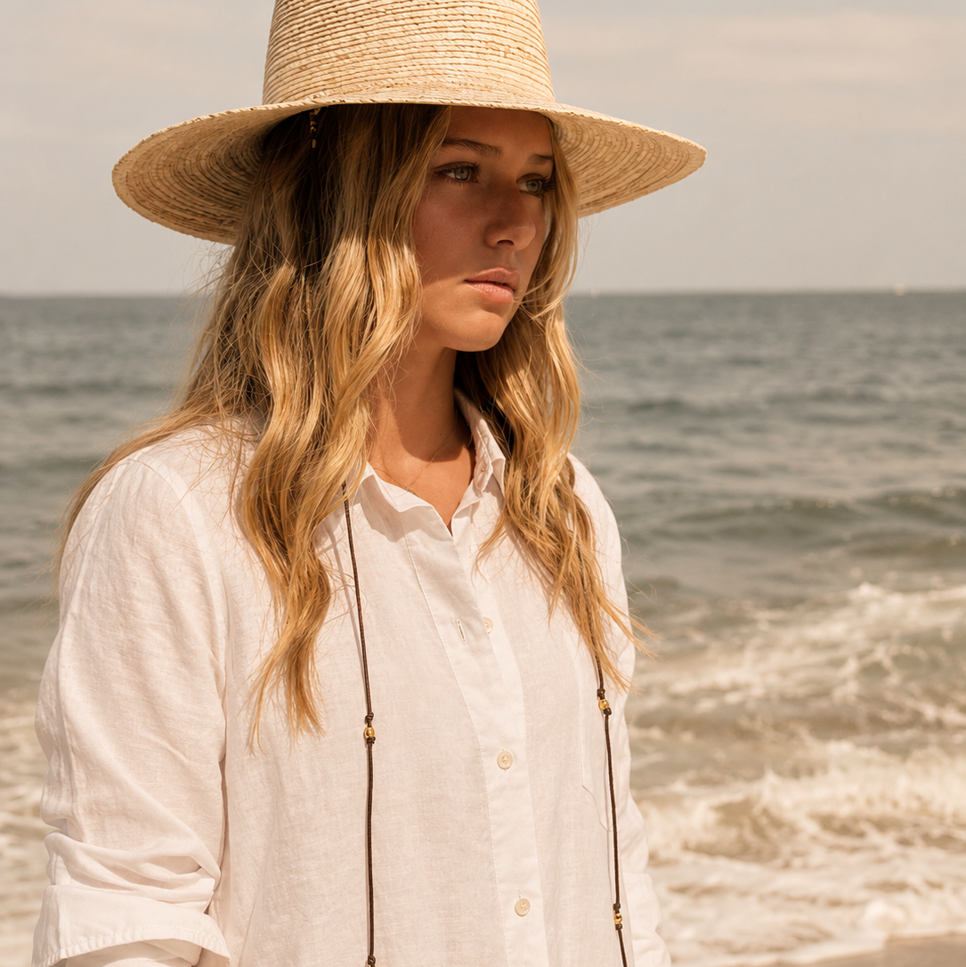A woman standing on a beach wearing a wide-brimmed straw hat, a white linen button-up shirt, and a long beaded necklace.