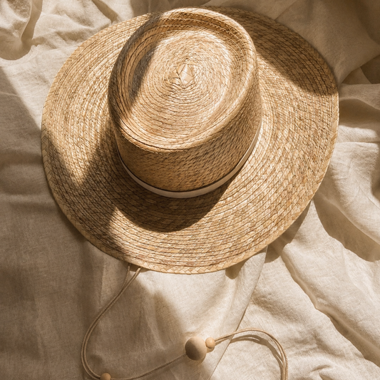 A natural straw wide-brim hat with a matching chin strap and wooden beads, styled on a textured beige linen fabric under warm, directional sunlight.