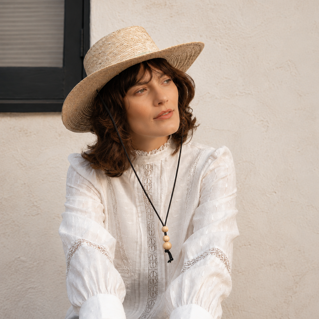 Woman wearing a wide-brimmed hat and white blouse against a plain wall.